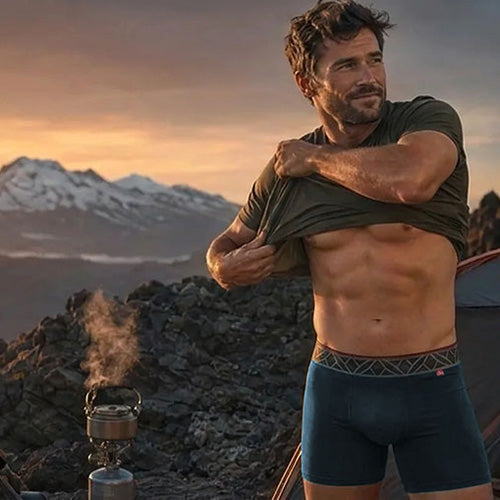A shirtless man in a mountain camping setting with a tent and stove, wearing an olive green t-shirt and Imperial Blue Anti-Gravity boxer briefs.